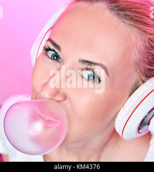 Fille blonde avec un casque d'exploser une bulle de gomme sur un fond rose Banque D'Images