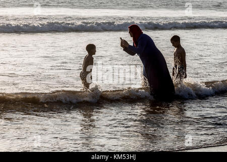 Agadir, Maroc, 22 octobre 2017 : femme musulmane entre dans l'océan atlantique dans sa robe pour prendre une photo de son fils dans la lumière du soir. Banque D'Images