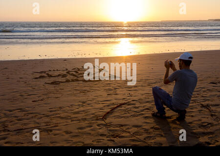 Agadir, Maroc, 22 octobre 2017 : un jeune homme de prendre une photo d'un signe d'amour qu'il a écrit avant sur la plage par l'océan atlantique. Banque D'Images