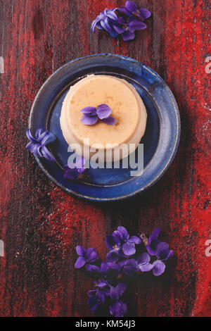 Vue de dessus sur la plaque avec pannacotta caramel servi avec des fleurs violettes sur fond de bois rouge Banque D'Images