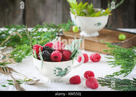 Vintage tasse de framboises et mûres servi avec thuja branches et vieux livre sur la table. Banque D'Images