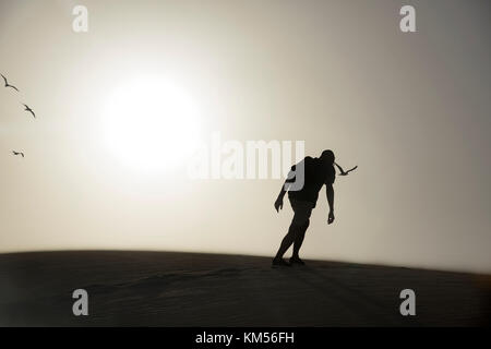 La silhouette d'un jeune homme de race blanche vu de dos portant un t-shirt et un short, et de transporter un sac à dos, la marche par le sable d'un désert, en ba Banque D'Images