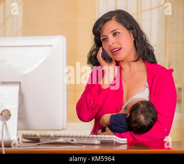 Belle jeune femme d'affaires travaillant dans le bureau alors qu'il était assis avec son petit garçon à son lieu de travail, l'allaitement pendant qu'elle est à l'aide de son portable dans un arrière-plan flou Banque D'Images