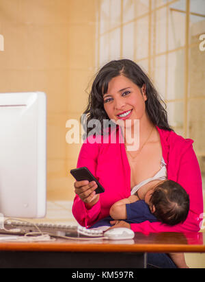 Belle jeune femme d'affaires travaillant dans le bureau alors qu'il était assis avec son petit garçon à son lieu de travail, l'allaitement pendant qu'elle est à l'aide de son portable dans un arrière-plan flou Banque D'Images