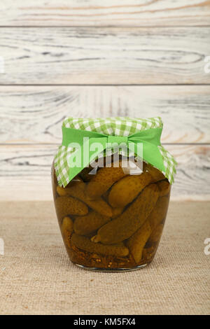 Close up d'un bocal en verre de petits concombres cornichons marinés , tissu à carreaux et décoration de porte de ruban vert sur toile over white woode Banque D'Images