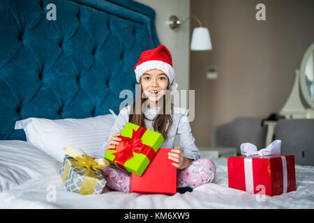 Belle petite fille à Santa's hat boîte cadeau d'ouverture, à la surprise alors qu'il était assis sur le lit chez lui Banque D'Images