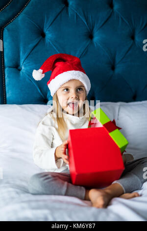 Surpris little girl in santa hat holding present alors qu'il était assis les jambes croisées au lit Banque D'Images