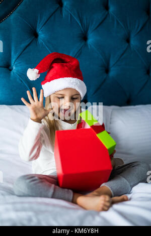 Surpris little girl in santa hat holding present alors qu'il était assis les jambes croisées au lit Banque D'Images