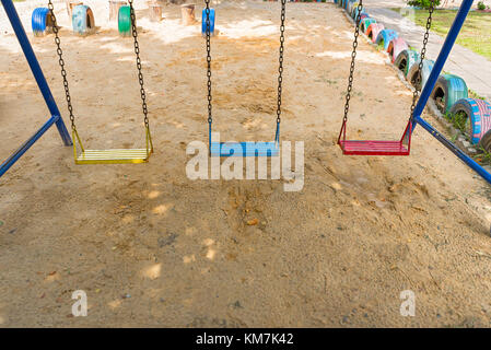 Trois vieux métal peint coloré des balançoires pour enfants dans une aire de jeu couverte de sable. Banque D'Images