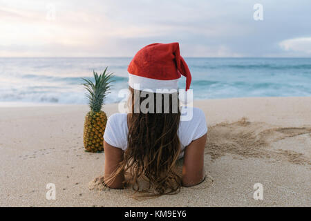 Femme portant un chapeau de Noël allongé sur la plage à côté d'un ananas, Haleiwa, Hawaii, États-Unis Banque D'Images