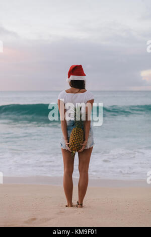 Femme portant un chapeau de Noël sur la plage tenant un ananas derrière son dos, Haleiwa, Hawaii, États-Unis Banque D'Images