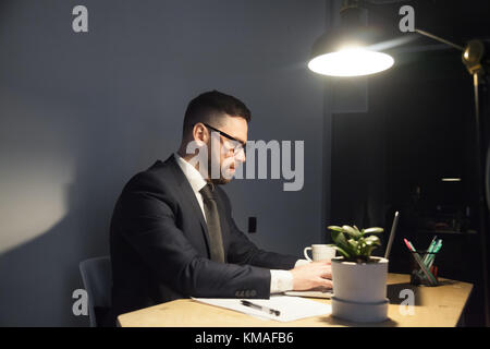Jeune ouvrier barbu en costume et cravate tapant sur ordinateur portable sous la lumière de la lampe. Gestionnaire moderne confiant essayant d'être à temps. Délai du travail, surmenage Banque D'Images
