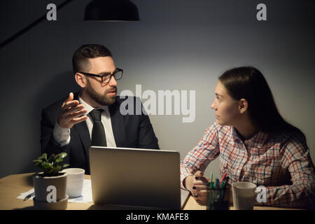 Mentor barbu à lunettes et costume expliquer jeune travailleuse des problèmes urgents de travail. travailler à échéance à la fin de nuit, overhours réunion. Banque D'Images