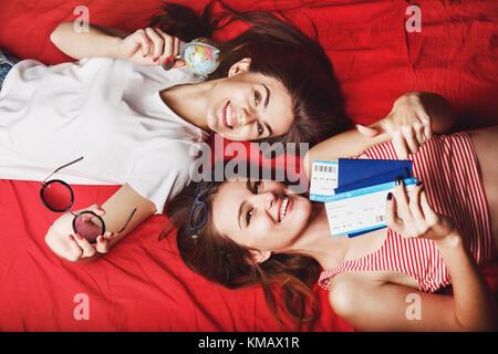 Deux cheerful friends girls holding les passeports, les billets d'avion et globe couché sur un lit rouge, intérieur travel concept Banque D'Images