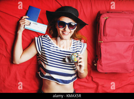 Cheerful girl holding passeport, billet d'avion et globe couché sur un lit rouge, intérieur travel concept Banque D'Images