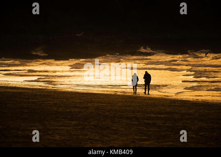 Coucher du soleil - un couple vu en silhouette marcher le long de la ligne de rivage au cours d'une intense coucher du soleil à la plage de Fistral Newquay Cornwall UK. Banque D'Images