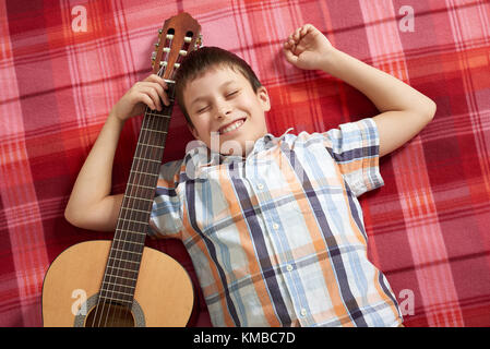 Garçon jouant de la musique à la guitare, se trouve sur une couverture à carreaux rouge, vue du dessus Banque D'Images