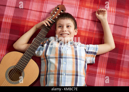 Garçon jouant de la musique à la guitare, se trouve sur une couverture à carreaux rouge, vue du dessus Banque D'Images