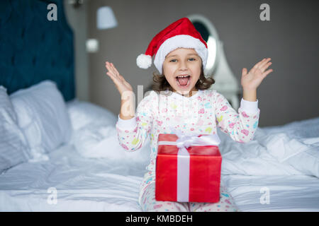 Surpris little girl in santa hat holding present alors qu'il était assis les jambes croisées au lit Banque D'Images