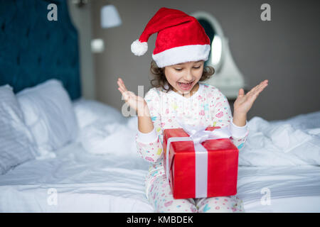 Surpris little girl in santa hat holding present alors qu'il était assis les jambes croisées au lit Banque D'Images