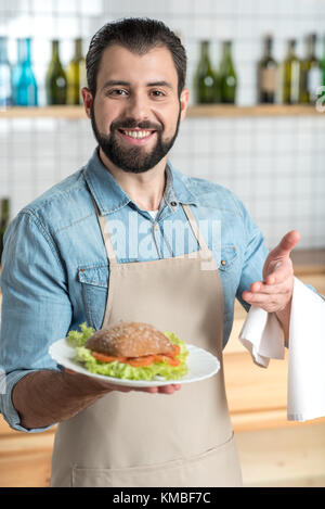 Friendly smiling waiter barbu tout en pointant sur le sandwich Banque D'Images