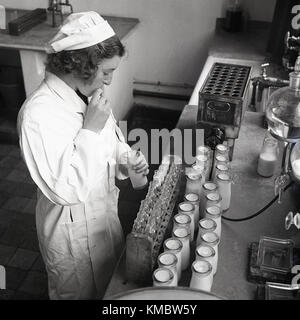 Années 1950, historiques, femme Technicien de laboratoire l'élaboration du lait d'une bouteille avec une paille pour faire tester des échantillons de tube pour contrôle. Banque D'Images