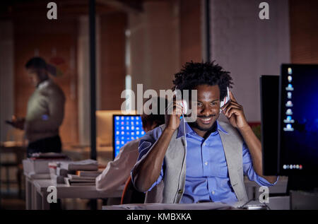 Smiling businessman with headphones listening to music,travailler tard à l'ordinateur de bureau sombre dans la nuit Banque D'Images