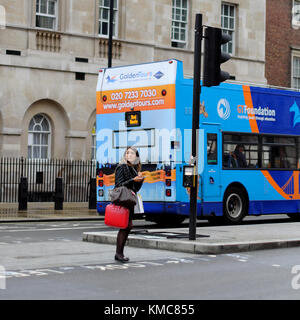 Brunette woman veste en cuir noire et portant deux sacs en cuir crossing road pendant l'utilisation de smartphone dans London, England, UK Banque D'Images