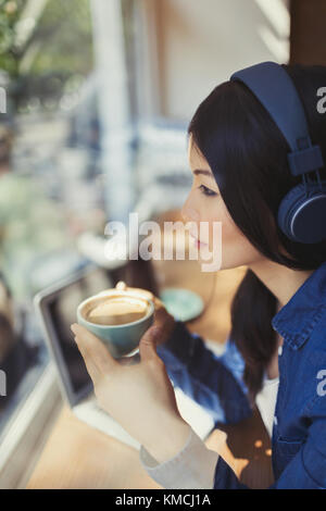 Jeune femme pensive buvant du café, écoutant de la musique avec des écouteurs à la fenêtre du café Banque D'Images