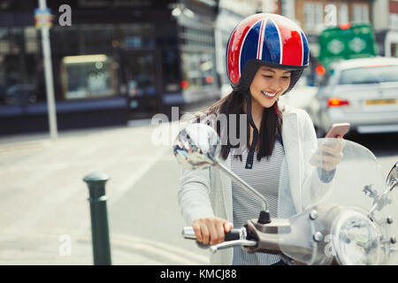 Jeune femme souriante envoyant un SMS avec un téléphone portable sur un scooter à moteur, portant un casque dans la rue urbaine Banque D'Images