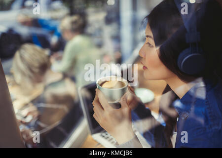 Jeune femme pensive à l'écoute de musique avec des écouteurs et à boire café à la fenêtre du café Banque D'Images