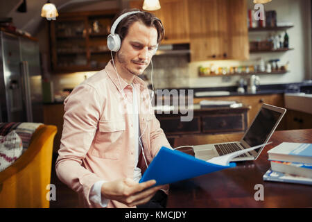 Homme avec un casque qui travaille à un ordinateur portable, lisant des documents dans la cuisine Banque D'Images