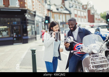 Jeune couple utilisant un téléphone cellulaire à moteur scooter sous le soleil rue urbaine Banque D'Images