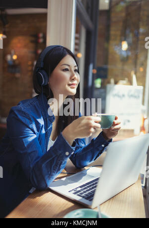 Jeune femme pensive à l'écoute de musique avec des écouteurs et à boire café à l'ordinateur portable dans la fenêtre du café Banque D'Images