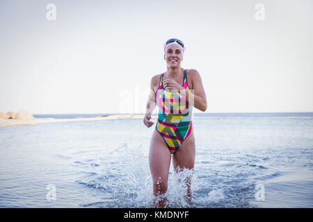 Portrait souriante, femme confiante, nageuse d'eau libre qui court et éclabousse dans les vagues de l'océan Banque D'Images