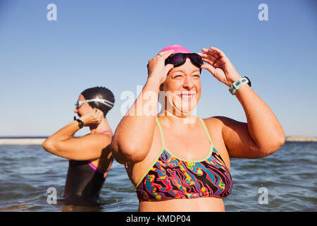 Souriante, heureuse femme nageuse en eau ouverte qui règle les lunettes de natation dans l'océan ensoleillé Banque D'Images