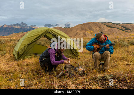 Petit-déjeuner de cuisson camping-femelle à mâle et réchaud de camping Camping-assis et jouant de la mandoline en face de tente,Jackson,Wyoming, USA Banque D'Images