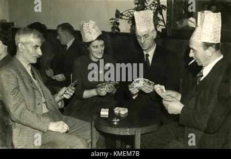 Années 1950, historiques, trois hommes et une femme assis ensemble autour d'une petite table de travail dans un club pour hommes jouant aux cartes et wearing party hats, England, UK. Banque D'Images