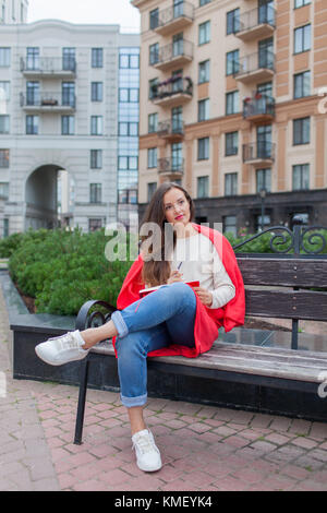 Une fille attrayante avec de longs cheveux bruns et un sourire blanc de dent s'assoit sur un banc et écrit ses pensées sur le fond urbain dans un carnet rouge.Elle Banque D'Images