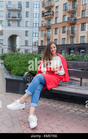 Une fille attrayante avec de longs cheveux bruns et un sourire blanc de dent s'assoit sur un banc et écrit ses pensées sur le fond urbain dans un carnet rouge.Elle Banque D'Images