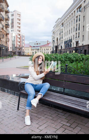 Une belle jeune femme dans un élégant hat est assis sur un banc dans un nouveau quartier et de lire un livre papier. Elle tourne les pages et sm Banque D'Images