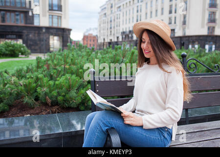 Une belle jeune femme dans un élégant hat est assis sur un banc dans un nouveau quartier et de lire un livre papier. Elle tourne les pages et sm Banque D'Images