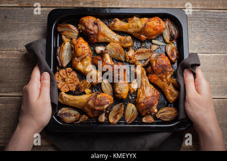 Mains de femmes tenant la plaque de cuisson chaude des pattes de poulet cuites au four avec des échalotes et de l'ail.Une femme met sur une table en bois sombre avec un plateau de frit Banque D'Images