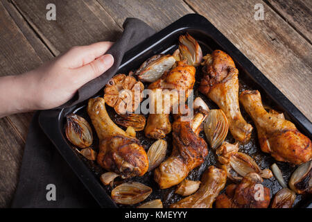 Mains de femmes tenant la plaque de cuisson chaude des pattes de poulet cuites au four avec des échalotes et de l'ail.Une femme met sur une table en bois sombre avec un plateau de frit Banque D'Images