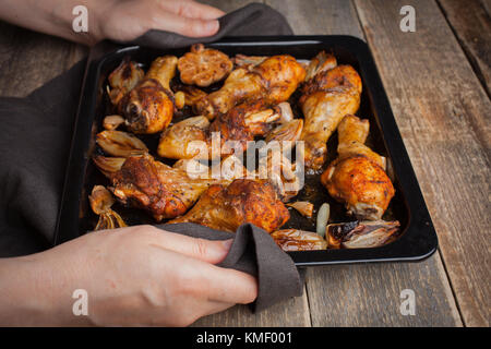 Mains de femmes tenant la plaque de cuisson chaude des pattes de poulet cuites au four avec des échalotes et de l'ail.Une femme met sur une table en bois sombre avec un plateau de frit Banque D'Images