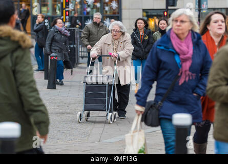 Personnes âgées femme marche dans une ville animée dans des magasins avec un chariot à roues dans le cadre d'un Walker, au Royaume-Uni. Banque D'Images