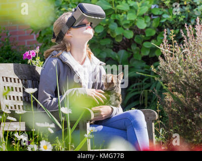 Jeune fille utilisant un casque de réalité virtuelle assis sur un banc de stationnement avec cat Banque D'Images