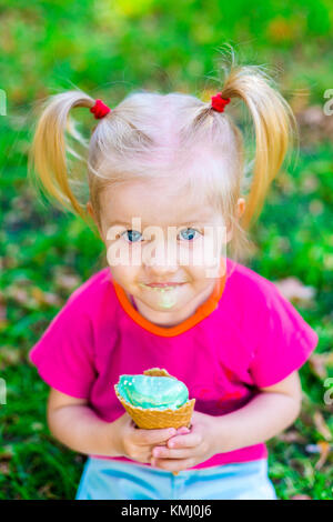 Peu drôle caucasian girl blonde aux yeux bleus avec deux queues sur la tête de manger une glace dans un cornet gaufré bleu tasse de assis sur l'herbe verte. tous faire face Banque D'Images