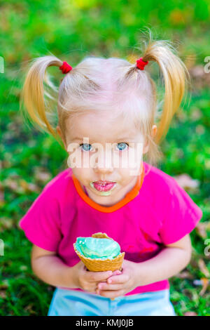 Peu drôle caucasian girl blonde aux yeux bleus avec deux queues sur la tête de manger une glace dans un cornet gaufré bleu tasse de assis sur l'herbe verte. tous faire face Banque D'Images