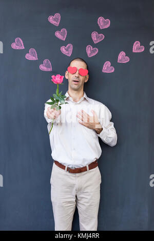 Seul adulte homme blanc avec des yeux en forme de coeur portant une chemise blanche et debout devant un tableau noir avec coeurs peints tenant une rose. Banque D'Images